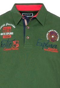 Grøn polo-shirt med røde farver ved kraven og broderede logoer. Har knaplukning, tekstureret stof og dekorative stikninger.