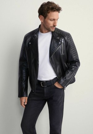 Mężczyzna w czarnej skórzanej kurtce motocyklowej, białej koszulce, czarnych dżinsach i czarnym pasku, patrzący w dół, z jedną ręką w kieszeni.