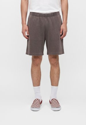 Mand iført grå knælange shorts, hvide crew-sokker og rødhvide ternede slip-on sko, stående mod en ensfarvet baggrund.