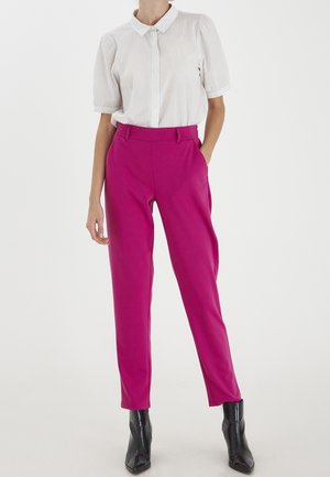 Pantalon rose ajusté, taille haute, avec une texture lisse. Associé à une chemise blanche à manches courtes, boutonnée, à rayures verticales.