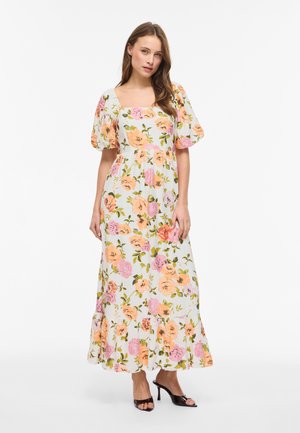 Nő fehér, virágos maxi ruhában, puffos ujjakkal és négyzet alakú nyakkivágással, fekete, magas sarkú szandállal párosítva, fehér háttér előtt áll.