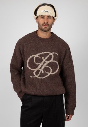 Homme portant un pull en maille marron avec un grand monogramme beige et un chapeau beige en peau lainée avec des oreillettes, sur un fond uni.