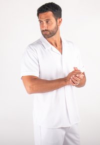 Chemise blanche à manches courtes, en tissu texturé, boutonnée, avec un col plat et une coupe décontractée. Portée avec un pantalon de couleur claire assorti.