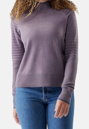 Kvinde iført en tætsiddende lilla sweater med ribstrikkede ærmer og kant, kombineret med blå jeans.