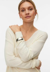 Femme aux cheveux blonds portant un pull en maille crème et un bracelet argenté brillant, les bras croisés et tenant doucement son épaule gauche.