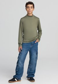 Długi top w oliwkowej zieleni z wysokim kołnierzem, zestawiony z luźnymi niebieskimi spodniami cargo z kieszeniami, noszony z czarnymi trampkami z beżowymi podeszwami.