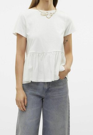 Femme portant un haut blanc à manches courtes avec des volants, un jean bleu clair et un collier abstrait doré, une main dans sa poche.