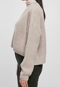 Pull en maille beige clair avec une texture épaisse et des épaules tombantes, doté d'un col arrondi et d'un ourlet raccourci, associé à un jean noir.