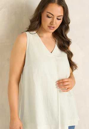 Kobieta o długich brązowych włosach, ubrana w bezrękawnik biały top, patrząca w dół, z jedną ręką delikatnie dotykającą tkaniny.