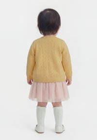 Tagurpidi seisva väikelapse seljas on kollane kootud kampsun, roosa tullekangast seelik, valged põlvesokid ja valged kingad, taust on ühtlane.