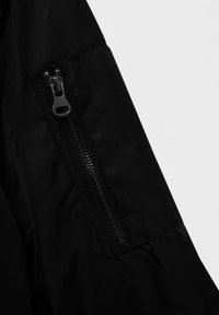 Manche d'une veste noire présentant une texture de tissu lisse, une poche latérale zippée avec une fermeture éclair noire, et des coutures renforcées.