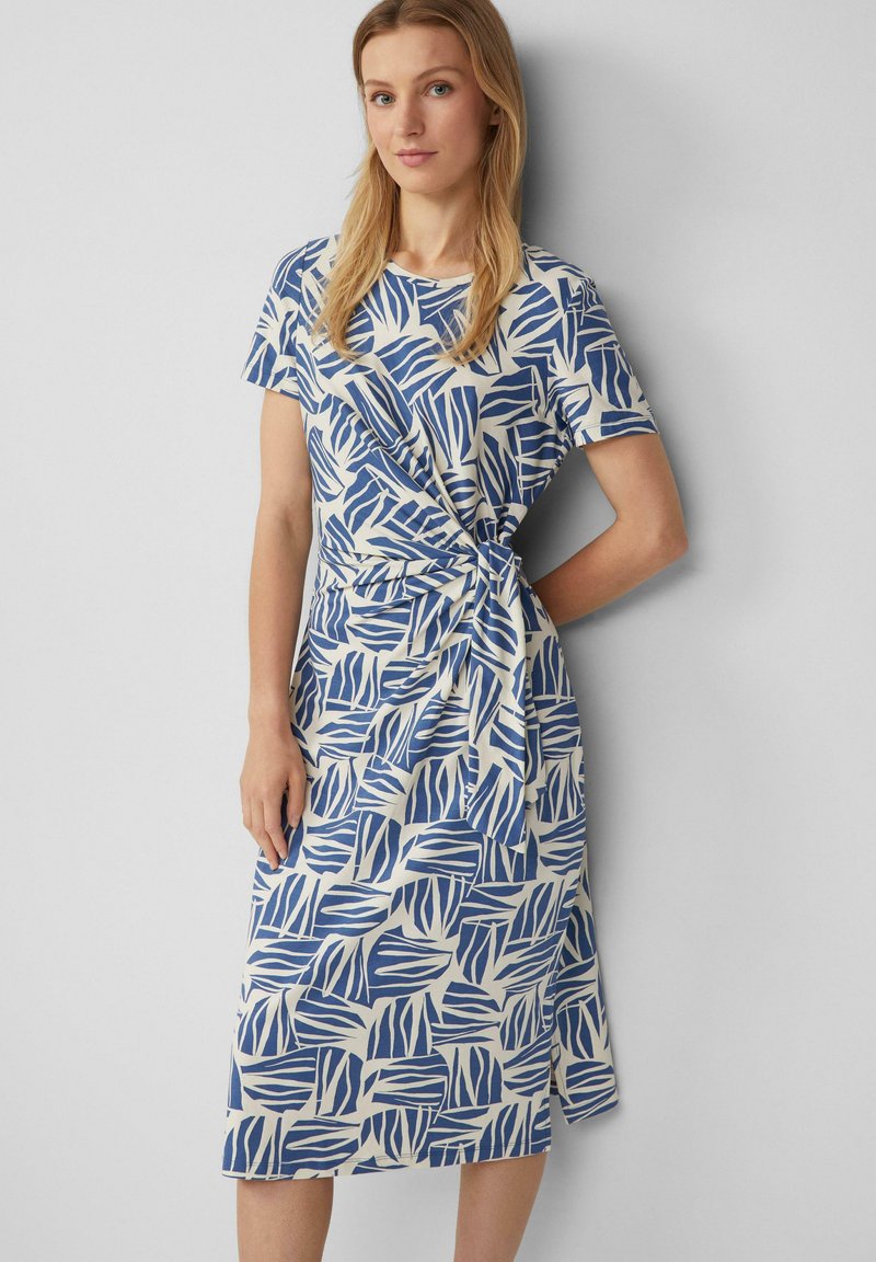 Niebieska i biała sukienka w wzory z krótkimi rękawami, z dekoltem kopertowym i paskiem w talii. Wykonana z gładkiego, lekkiego materiału.