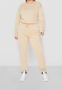 Frau in beigem, langärmligem Cropped-Top und passenden Jogginghosen mit weißen High-Top-Sneakern, die vor einem schlichten Hintergrund steht.