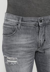 Jeans de mezclilla gris con un acabado deslavado, diseño de cinco bolsillos y un desgarro en la parte superior del muslo izquierdo. Cierre con botón metálico.