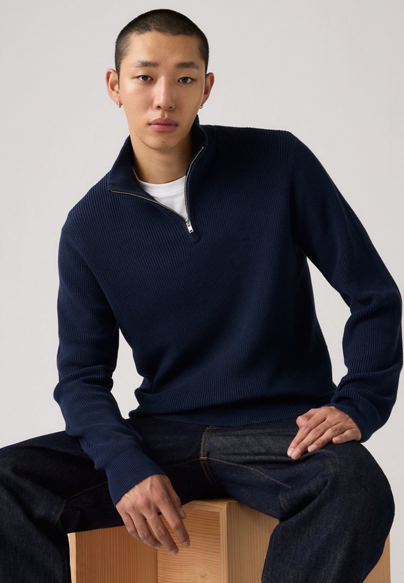 Marineblauer gerippter Pullover mit einem Viertel-Reißverschluss am Kragen, getragen über einem weißen Hemd. Modell sitzt auf einem Holzblock, kombiniert mit dunklen Denim-Jeans.