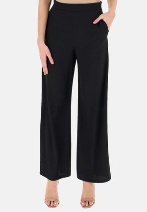 Pantalon noir à jambes larges avec poches, porté par une personne debout, associé à des sandales beige à bouts ouverts sur un fond uni.