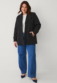 Zwarte trenchcoat met brede kraag, knoopsluiting en zijzakken, gecombineerd met blauwe broek met wijde pijpen en puntige zwarte hakken.