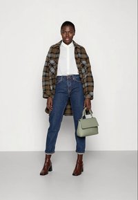 Cappotto a quadri marrone e nero, camicia bianca con colletto, jeans skinny blu, stivali marroni con tacco, in mano una borsa leggera verde.