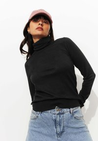 Une femme portant une casquette rose, un pull à col roulé noir et un jean bleu clair se tient contre un fond blanc, les mains derrière le dos.