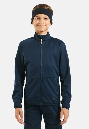 Ein Junge steht, trägt eine marineblaue Zip-up-Jacke, passende Hosen und ein schwarzes Stirnband, und lächelt vor einem einfarbigen hellen Hintergrund.