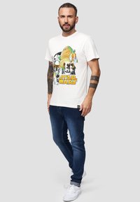 Weißes T-Shirt mit einem bunten "Star Wars"-Grafik, kombiniert mit dunkelblauen Jeans und weißen Sneakers. Einfaches Design mit kurzen Ärmeln.