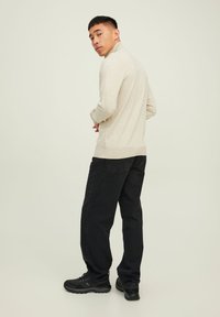 Suéter beige de manga larga con un cuello alto, combinado con jeans negros de corte suelto y zapatillas negras, mostrado desde atrás contra un fondo claro.