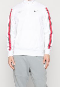 Vit zip-up sweatshirt med röda sidostråk och Nike-logotyper, tillverkad av mjukt material, i kombination med grå byxor.