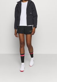 Black hooded jacket, white T-shirt, black shorts with neon green logo, knee-high black socks with pink accents, and white soccer shoes.