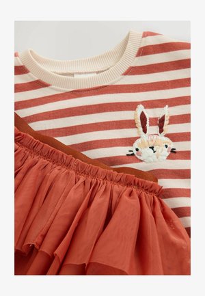 Maglietta a righe rosse e bianche con un coniglio ricamato. Presenta un sovrapposto trasparente con volant in terracotta nella parte inferiore e un colletto a coste.