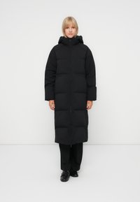 Manteau long noir en duvet avec fermeture éclair, capuche et grandes sections créant une texture matelassée. Il possède des manches larges et une coupe droite.