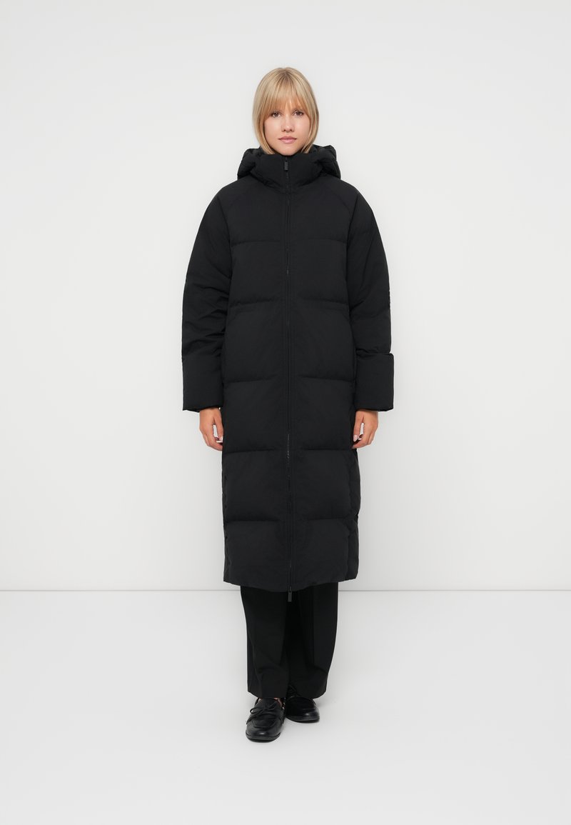 Manteau long noir en duvet avec fermeture éclair, capuche et grandes sections créant une texture matelassée. Il possède des manches larges et une coupe droite.