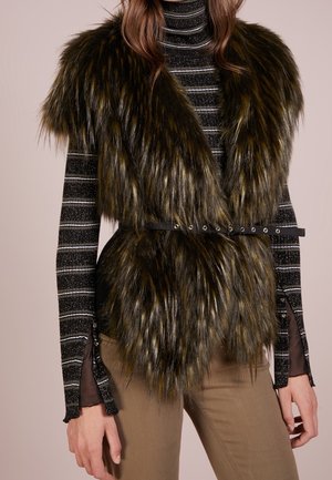 Kobieta ubrana w ciemnobrązową kamizelkę bez rękawów z futerka ekologicznego z czarnym paskiem, nałożoną na czarny i srebrny pasiasty golf oraz beżowe spodnie.