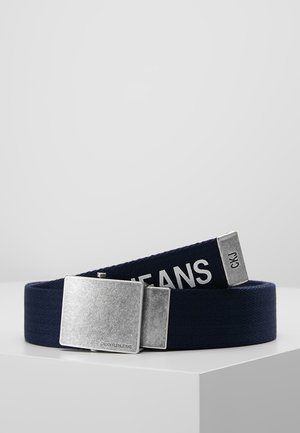 Ceinture en tissu marine enroulée sur une surface blanche avec une grande boucle rectangulaire argentée gravée "CALVIN KLEIN JEANS" et "CKJ" au bout de la ceinture.