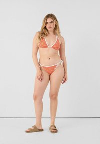 Ensemble de bikini à motif orange avec une bordure blanche, comprenant un haut triangulaire et un bas à nouer sur les côtés, associé à des sandales à glissière marron.
