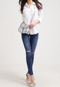 Kobieta ubrana w białą koszulę, podarte obcisłe jeansy, srebrne szpilki oraz niosąca srebrną torebkę na ramię z czarnymi detalami szwu.