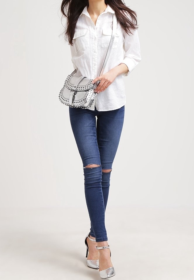 Kobieta ubrana w białą koszulę, podarte obcisłe jeansy, srebrne szpilki oraz niosąca srebrną torebkę na ramię z czarnymi detalami szwu.