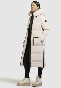 Beige puffer coat with a high collar, zipper, and front pockets. Worn over a black outfit and paired with beige chunky boots.