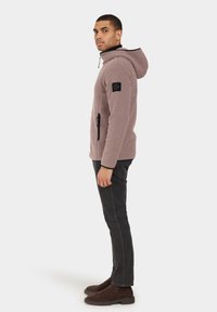 Sudadera de forro polar en rosa claro con cremallera y dos bolsillos; presenta un parche de logo negro en la manga y una capucha para mayor calidez.