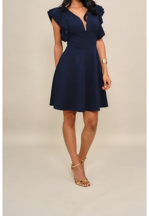 Femme portant une robe sans manches bleu marine avec des épaules à volants, un décolleté en V profond et des sandales à talons hauts dorées, debout devant un fond uni.