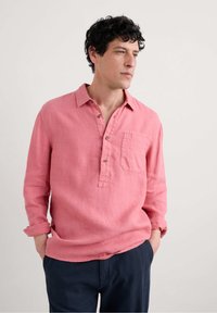 Långärmad, rosa linneskjorta med krage, knappslå och bröstficka, bärs avslappnat med marinblå byxor.