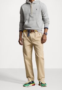 Felpa grigia con cappuccio e coulisse, chinos beige e sneakers multicolori con dettagli verdi, blu e rossi. Visibili una cintura marrone e dettagli di un orologio.