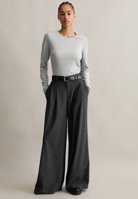 Szürke, hosszú ujjas póló, amelyet magas derekú, bő szárú fekete nadrággal párosítanak, fekete övvel aranycsattal és mély oldalsó zsebekkel.