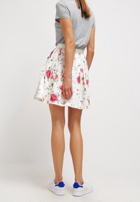 Blumiges A-Linien-Rock mit einem weißen Hintergrund, der rosa Rosen und grüne Blätter zeigt. Kombiniert mit einem grauen T-Shirt und weißen Sneakers mit blauen Akzenten.