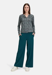 Langärmlige Bluse mit einem schwarz-türkisen geometrischen Muster, kombiniert mit türkisfarbenen weiten Hosen und weißen Sneakers.