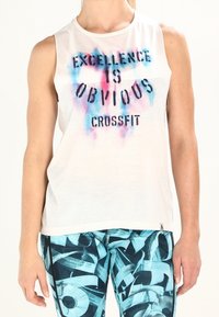 Osoba ubrana w białą bluzkę bez rękawów z napisem "Excellence is Obvious CrossFit" oraz niebieskie legginsy sportowe w wzory.