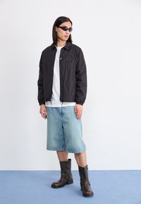 Black lightweight jacket with a logo, white t-shirt, loose denim shorts, and brown ankle boots with a buckle detail. Set against a light background.