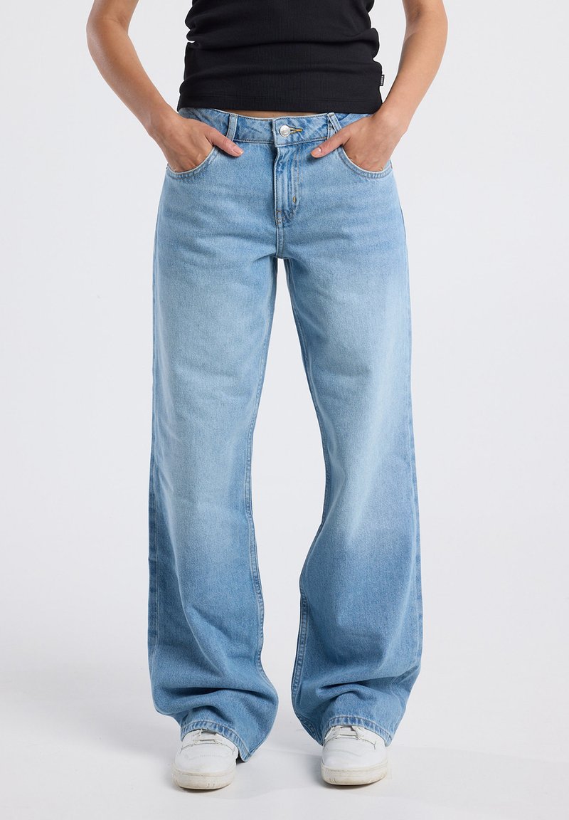 Person wearing light blue wide-leg jeans with hands in pockets and white sneakers, standing against a plain background.