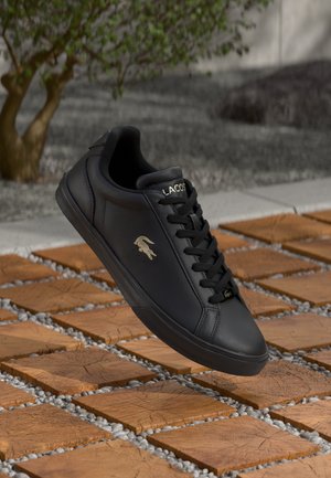 Black leather sneaker with gold "LACOSTE" branding and iconic crocodile logo. Features flat laces and a rubber sole, set against wooden tiles.