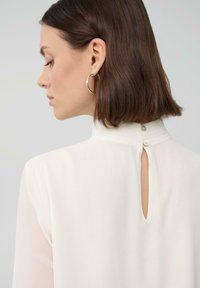 Woman with short brown hair wearing silver hoop earring and white high-collar blouse buttoned at the back with a keyhole opening.