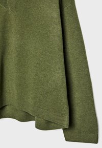 Groene gebreide trui met ribbelmanchetten en onderrand, textuur van de stof zichtbaar, hangend tegen een eenvoudige lichte achtergrond.